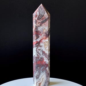 May include: A tall, rectangular crystal obelisk with a pointed top. The stone features a complex pattern of red, white, gray, and tan swirls and veins. The obelisk is standing upright on a white surface against a black background.