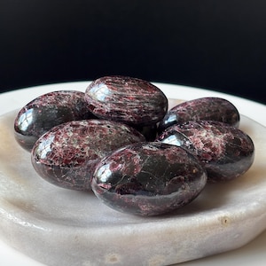 May include: A collection of polished, oval-shaped garnet stones. The stones are a deep burgundy color with visible internal patterns and are arranged in a white, shallow dish. The background is a dark, solid color.