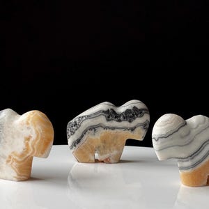 May include: Three carved stone animal figurines shaped like bison, each featuring distinct banded patterns in shades of white, gray, and amber. The smooth, polished surfaces display natural mineral layers and textures throughout the solid forms.