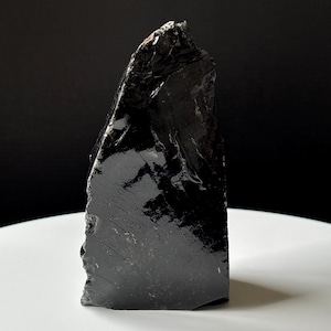 May include: A large, jagged piece of black obsidian with a highly reflective, glass-like surface. The stone features sharp, irregular edges and a smooth, glossy texture that catches the light, showcasing its deep, dark, and opaque coloration.