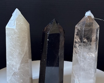 May include: Three crystal obelisks. One is a dark, opaque black. The other two are translucent, one a light smoky gray and the other clear. Each has a pointed top and a faceted, rectangular body. They stand upright against a dark background.