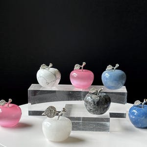 May include: A collection of decorative apple sculptures in various colors, including pink, blue, white, and gray. Each apple features a silver-colored stem and leaf detail. The apples are displayed on clear acrylic blocks against a black background.