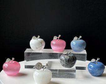 May include: A collection of decorative apple sculptures in various colors, including pink, blue, white, and gray. Each apple features a silver-colored stem and leaf detail. The apples are displayed on clear acrylic blocks against a black background.