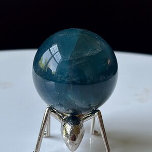 Puede incluir: Una esfera de cristal de color verde azulado se encuentra sobre un soporte de metal plateado. La esfera tiene una superficie lisa y reflectante. El soporte est&aacute; dise&ntilde;ado con una forma &uacute;nica de hormiga, con cabeza y patas. El fondo es una superficie blanca con un fondo negro.