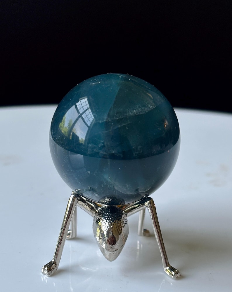 Puede incluir: Una esfera de cristal de color verde azulado se encuentra sobre un soporte de metal plateado. La esfera tiene una superficie lisa y reflectante. El soporte est&aacute; dise&ntilde;ado con una forma &uacute;nica de hormiga, con cabeza y patas. El fondo es una superficie blanca con un fondo negro.