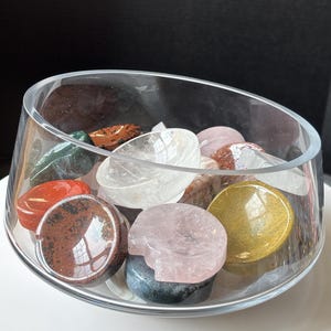 Puede incluir: Un cuenco de vidrio transparente lleno de varias piedras preciosas pulidas. Las piedras son de color rojo, verde, marrón, rosa, amarillo y transparente. El cuenco está sobre una superficie blanca.