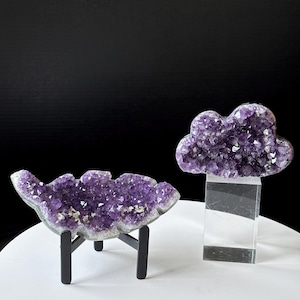 May include: Two amethyst crystal sculptures. One is a cloud-shaped cluster on a clear acrylic stand. The other is a larger, cloud-shaped cluster on a black metal stand. Both feature deep purple crystal formations.