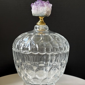 Könnte beinhalten: Ein klares Glasgefäß mit dekorativem Deckel, gekrönt von einem Cluster aus lila Amethystkristallen. Das Gefäß hat eine runde Form und eine strukturierte, gemusterte Oberfläche. Der Deckel ist mit einem goldfarbenen Metallstück befestigt. Das Gefäß wird auf einem transparenten, rechteckigen Sockel präsentiert.