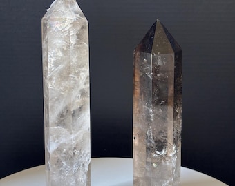 May include: Two quartz crystal towers. One is clear with a white tip, the other is a darker smoky quartz. Both are rectangular with pointed tops. The crystals are on a white surface with a dark background.