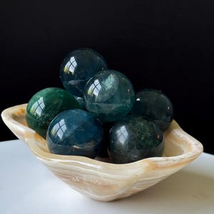 Puede incluir: Una colecci&oacute;n de piedras preciosas esf&eacute;ricas pulidas, en tonos verdes y azules, dispuestas en un cuenco decorativo de color crema. El cuenco tiene un aspecto veteado natural. Las piedras son lisas y reflectantes.