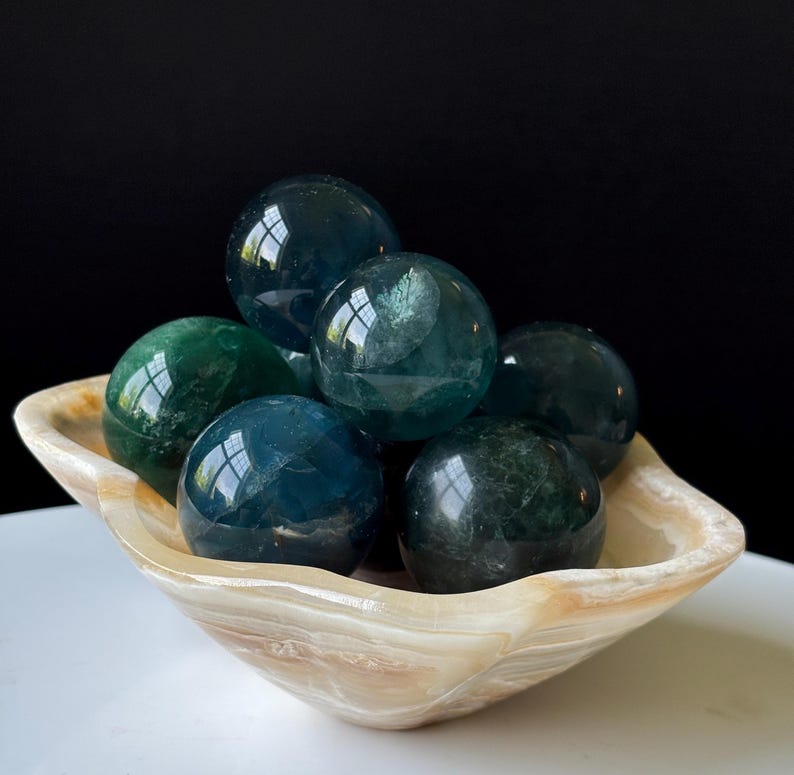 Puede incluir: Una colecci&oacute;n de piedras preciosas esf&eacute;ricas pulidas, en tonos verdes y azules, dispuestas en un cuenco decorativo de color crema. El cuenco tiene un aspecto veteado natural. Las piedras son lisas y reflectantes.