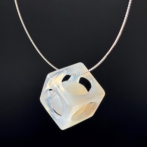 Puede incluir: Un collar con colgante translúcido en forma de cubo con recortes redondeados. El colgante cuelga de una delicada cadena de plata. El cubo tiene una calidad nacarada y opalescente, sobre un fondo negro.