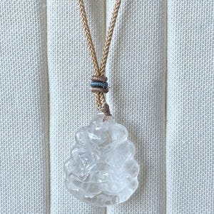 May include: A clear, carved pendant necklace with a beige cord. The pendant is a detailed, translucent figure. The cord is knotted with a small, decorative accent. The background is a textured, off-white fabric.