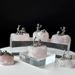 May include: Several silver miner figurines are displayed on light pink quartz crystal formations. The figurines are detailed, and the quartz pieces vary in size and shape. The quartz is displayed on clear acrylic blocks, creating a unique display.