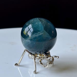 Puede incluir: Una esfera de cristal de color verde azulado se encuentra sobre un soporte met&aacute;lico plateado con forma de insecto. La esfera refleja el entorno. El soporte tiene seis patas y dos antenas.