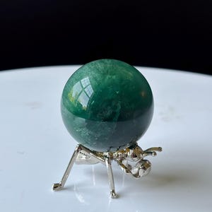 Puede incluir: Una esfera de cristal verde pulido, apoyada sobre un soporte de metal plateado. La esfera refleja la luz, mostrando su superficie lisa. El soporte tiene un dise&ntilde;o de insecto estilizado &uacute;nico, agregando un toque art&iacute;stico a la exhibici&oacute;n.