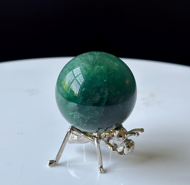 Puede incluir: Una esfera de cristal verde pulido, apoyada sobre un soporte de metal plateado. La esfera refleja la luz, mostrando su superficie lisa. El soporte tiene un dise&ntilde;o de insecto estilizado &uacute;nico, agregando un toque art&iacute;stico a la exhibici&oacute;n.