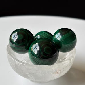 Può includere: Quattro sfere di malachite levigate con motivi a bande concentriche verde scuro sono disposte all'interno di una piccola ciotola di vetro trasparente e strutturato. Le sfere presentano una superficie liscia e riflettente con strati circolari.
