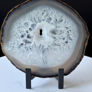 Pode incluir: Uma fatia de ágata polida, mostrando faixas concêntricas de cinza, branco e castanho. A pedra é montada em um suporte de metal preto, revelando intrincadas formações cristalinas e uma cavidade central. A pedra natural é um item decorativo.