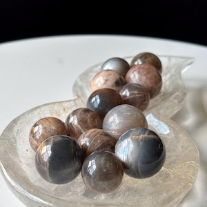 May include: A collection of polished, spherical gemstones in shades of brown, gray, and tan. The stones are displayed in a clear, leaf-shaped dish. The gemstones have a reflective, glossy surface.
