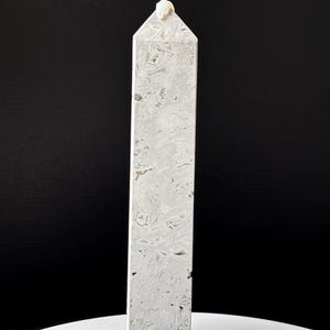 May include: A tall, rectangular, light gray stone obelisk with a pointed top. The obelisk has a textured surface with a pattern of small, irregular shapes. A small, light-colored stone is affixed to the top. The obelisk is displayed on a white surface against a black background.