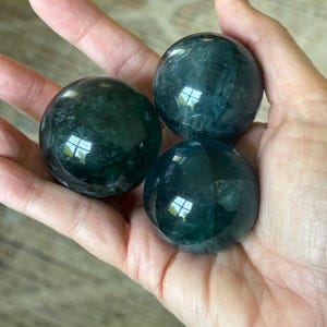 Puede incluir: Tres esferas de fluorita pulida, de color verde oscuro, se sostienen en una mano. Las esferas tienen una superficie lisa y reflectante, con sutiles variaciones de color y patrones internos. Es un art&iacute;culo decorativo natural.