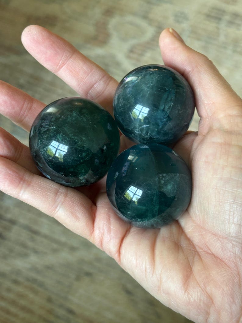 Puede incluir: Tres esferas de fluorita pulida, de color verde oscuro, se sostienen en una mano. Las esferas tienen una superficie lisa y reflectante, con sutiles variaciones de color y patrones internos. Es un art&iacute;culo decorativo natural.