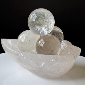 May include: A clear, leaf-shaped bowl carved from stone holds four smooth, spherical stones. Three of the spheres are transparent and colorless, while one sphere is a translucent, smoky gray color. The bowl has a textured, crystalline exterior.