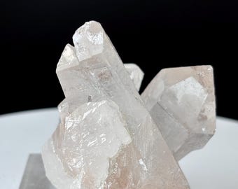 May include: A cluster of clear quartz crystals with a pale pink hue. The crystals are sharply pointed and arranged in a layered formation. The background is black, highlighting the translucent quality of the quartz.