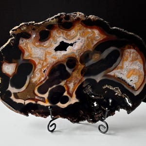 May include: A polished agate slice featuring a complex pattern of deep black, translucent amber, and creamy white bands. The stone has a jagged, natural outer edge and is displayed on a black metal wire stand with two curved support arms.