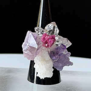 May include: A ring featuring a cluster of raw crystals including clear quartz, light purple amethyst, a bright magenta mineral formation, and a textured white crystalline structure, all arranged together on a dark, conical display stand.