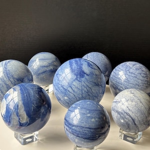 May include: A collection of blue quartz spheres with white veining, displayed on clear acrylic stands. The polished gemstones vary in size and reflect light, creating a visually appealing arrangement. The spheres are a mix of light and dark blue.