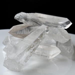 May include: A cluster of clear, translucent quartz crystal points with sharp, naturally faceted edges. The crystals are stacked together, displaying varying degrees of transparency, internal fractures, and smooth, reflective surfaces.