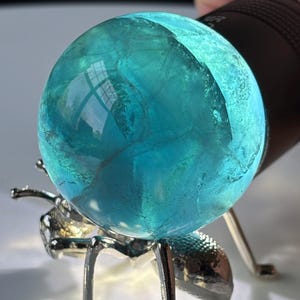 Puede incluir: Una bola de cristal transl&uacute;cida, de color turquesa, se muestra sobre un soporte plateado con forma de hormiga. La bola de cristal est&aacute; iluminada por una linterna negra, creando un efecto brillante. El soporte tiene seis patas y dos antenas.