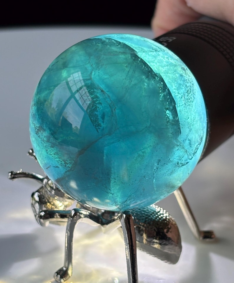 Puede incluir: Una bola de cristal transl&uacute;cida, de color turquesa, se muestra sobre un soporte plateado con forma de hormiga. La bola de cristal est&aacute; iluminada por una linterna negra, creando un efecto brillante. El soporte tiene seis patas y dos antenas.
