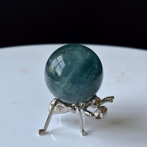 Puede incluir: Una esfera de cristal de color verde azulado exhibida sobre un soporte met&aacute;lico plateado con forma de hormiga. La esfera tiene una superficie lisa y pulida. El soporte tiene seis patas y un dise&ntilde;o detallado de hormiga. El fondo es una superficie blanca borrosa.