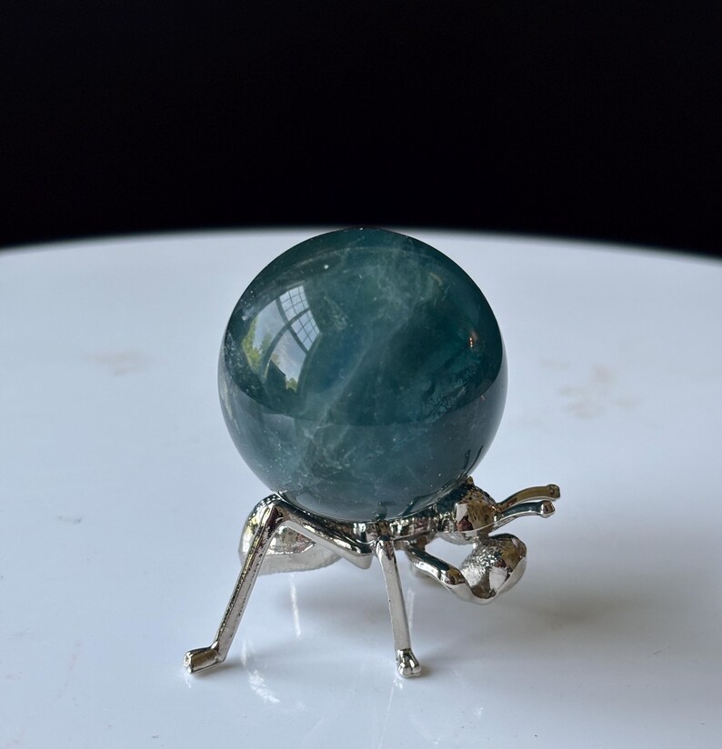 Puede incluir: Una esfera de cristal de color verde azulado exhibida sobre un soporte met&aacute;lico plateado con forma de hormiga. La esfera tiene una superficie lisa y pulida. El soporte tiene seis patas y un dise&ntilde;o detallado de hormiga. El fondo es una superficie blanca borrosa.