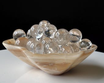 May include: A collection of clear, spherical quartz crystal balls piled inside a shallow, scalloped bowl made of polished, banded beige and cream-colored stone. The smooth, translucent spheres vary slightly in size and reflect light on their surfaces.
