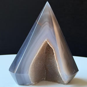 Puede incluir: Una escultura de piedra de ágata pulida con forma de pirámide, con un exterior gris translúcido y liso que presenta sutiles bandas blancas. La sección frontal está tallada para revelar un interior de geoda cristalina texturizada en tono marrón claro.