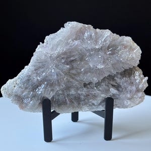 May include: A raw, translucent amethyst crystal cluster with a pale lavender hue and jagged, crystalline formations. The specimen is displayed upright on a black metal stand, showcasing its natural, irregular shape and multifaceted, light-reflecting surface.