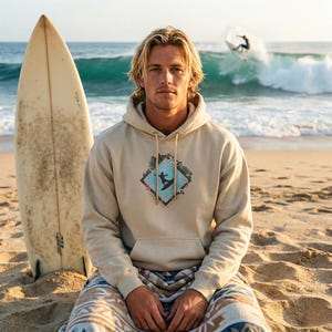 Könnte beinhalten: Beigefarbener Kapuzenpullover mit einem rautenförmigen Aufdruck eines Surfers auf einer Welle. Ein Surfbrett lehnt an der Person. Die Person sitzt an einem Sandstrand mit Meereswellen im Hintergrund. Der Hoodie hat eine Vordertasche und eine Kapuze mit Kordelzug.