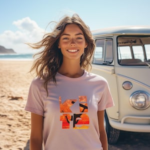 Puede incluir: Una mujer lleva una camiseta rosa con un gráfico de una puesta de sol y una silueta de una persona haciendo kitesurf. El gráfico está en tonos de naranja, amarillo y rojo. El texto "KITE" está superpuesto al gráfico. La mujer está de pie frente a una furgoneta blanca.