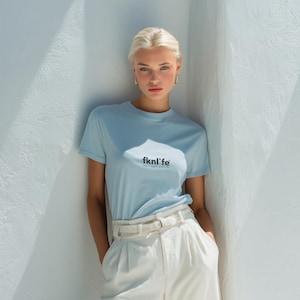 Puede incluir: Una camiseta azul claro con el texto "fknl'fe" en negro, combinada con pantalones blancos y un cinturón blanco. La modelo se apoya en una pared blanca, creando un look casual y elegante.