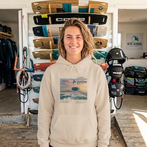 Puede incluir: Una sudadera con capucha beige con un gráfico frontal con el texto "TARIFA KITESURFING PARADISE" y una imagen del océano. La sudadera con capucha la lleva una persona de pie frente a una exhibición de tablas de surf y equipo de kitesurf.