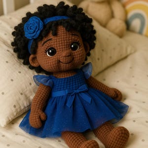 May include: A handmade crochet doll with dark skin, black curly hair, and a blue headband with a flower. The doll wears a blue dress with a bow and is lying on a bed with a teddy bear in the background.