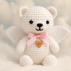 May include: A white crocheted teddy bear with angel wings, a pink bow, and a wooden heart. The bear has black eyes and a black nose. Text on the image reads "Heavenly hugs await..." and "www.crochix.com".