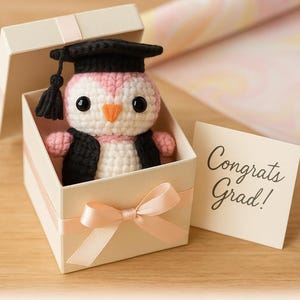 May include: A handmade crochet penguin wearing a graduation cap and gown, presented in a gift box tied with a peach ribbon. The penguin is white, pink, and black. A card reads "Congrats Grad!" and the text "Crafted with care, gifted with love."