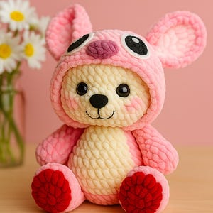 May include: A handmade pink and cream-coloured crochet teddy bear wearing a pink hood with black eyes and a black nose. The bear has red feet and is sitting on a wooden surface. Text reads: Make a meaningful gift for someone who means the world to You.
