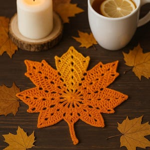 May include: A handmade orange and yellow crocheted maple leaf coaster on a dark wooden table, with a lit candle and a cup of tea with lemon. Dried leaves are arranged around the coaster, creating an autumnal scene.