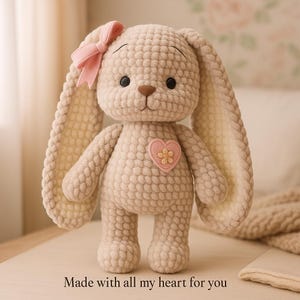 以下が含まれることがあります： ピンクのリボンと長い耳が付いた、手作りのベージュのかぎ針編みのウサギ。黒い目、茶色の鼻、黄色の花が付いたピンクのハート型のパッチが付いています。下部のテキストは「Made with all my heart for you」と書かれています。ぬいぐるみ。